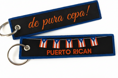 Two PUERTO RICAN luggage tags with blue and white embroidery, one with the text 'de pura cepa!' and the other displaying the Puerto Rican flag. Both tags have a keyring attachment.