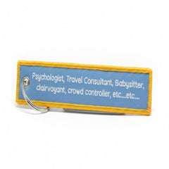 A rectangular embroidered patch with a light blue background and yellow border, featuring white embroidered text listing job titles like psychologist, travel consultant, and babysitter. A silver metal split ring is attached on the left side, set against a solid blue background.