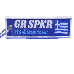 A rectangular embroidered keychain patch with a blue background and white border, featuring the text 'GR SPKR it's all Greek to me!' alongside a Greek flag design. A silver metal key ring is attached on the left.